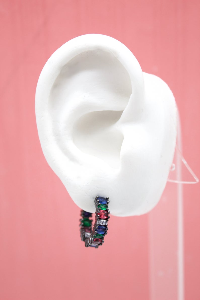 A white mannequin ear displays a black metal hoop earring encrusted with multicolored cubic zirconia stones.