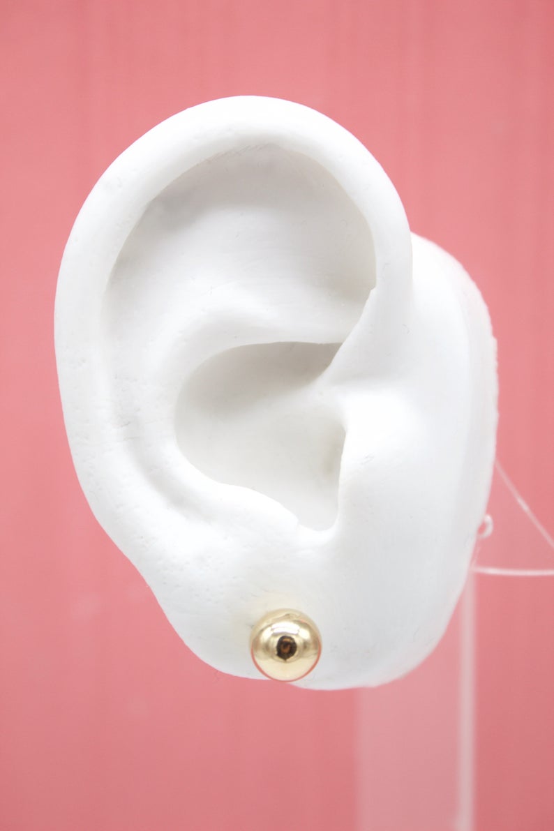 A white mannequin ear displays a polished gold ball stud earring against a pink background.