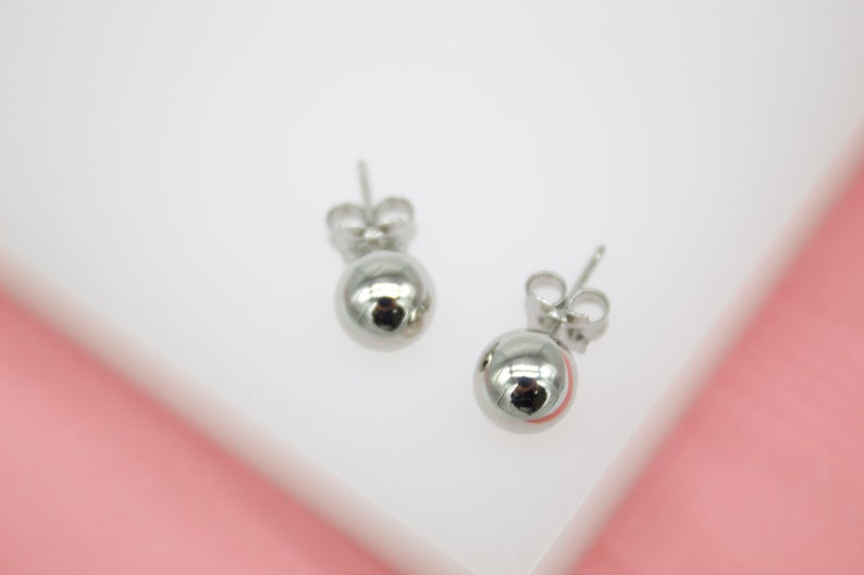 A pair of polished silver ball stud earrings rests against a white and pink background.