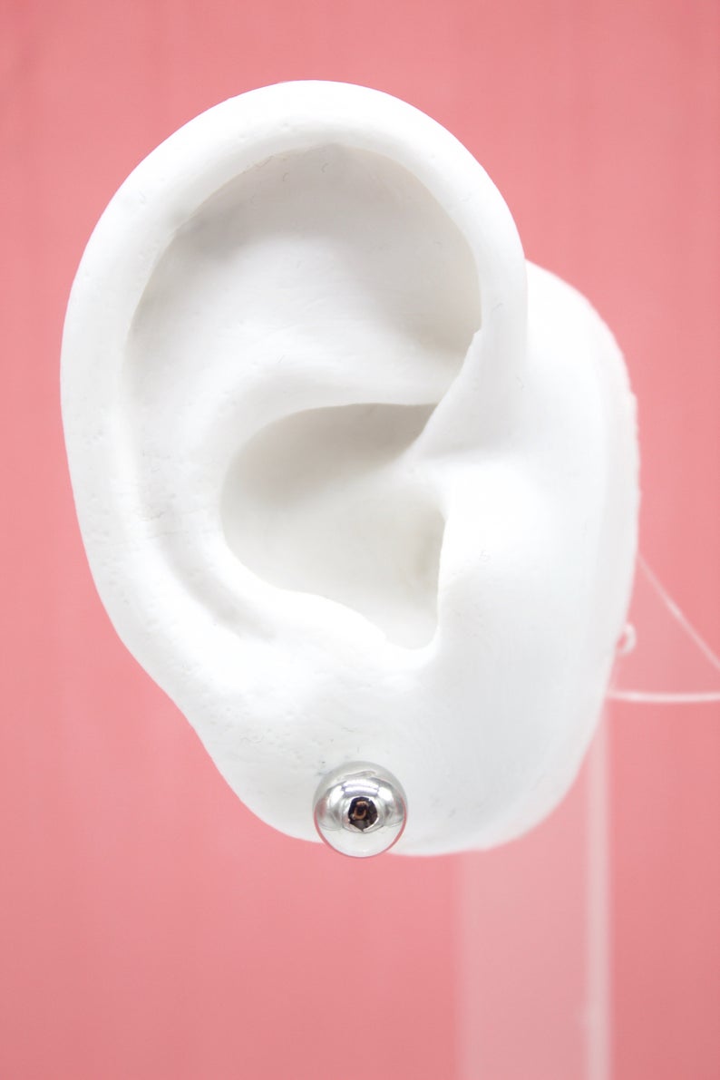 A white model ear displays a shiny silver ball stud earring against a soft pink background.