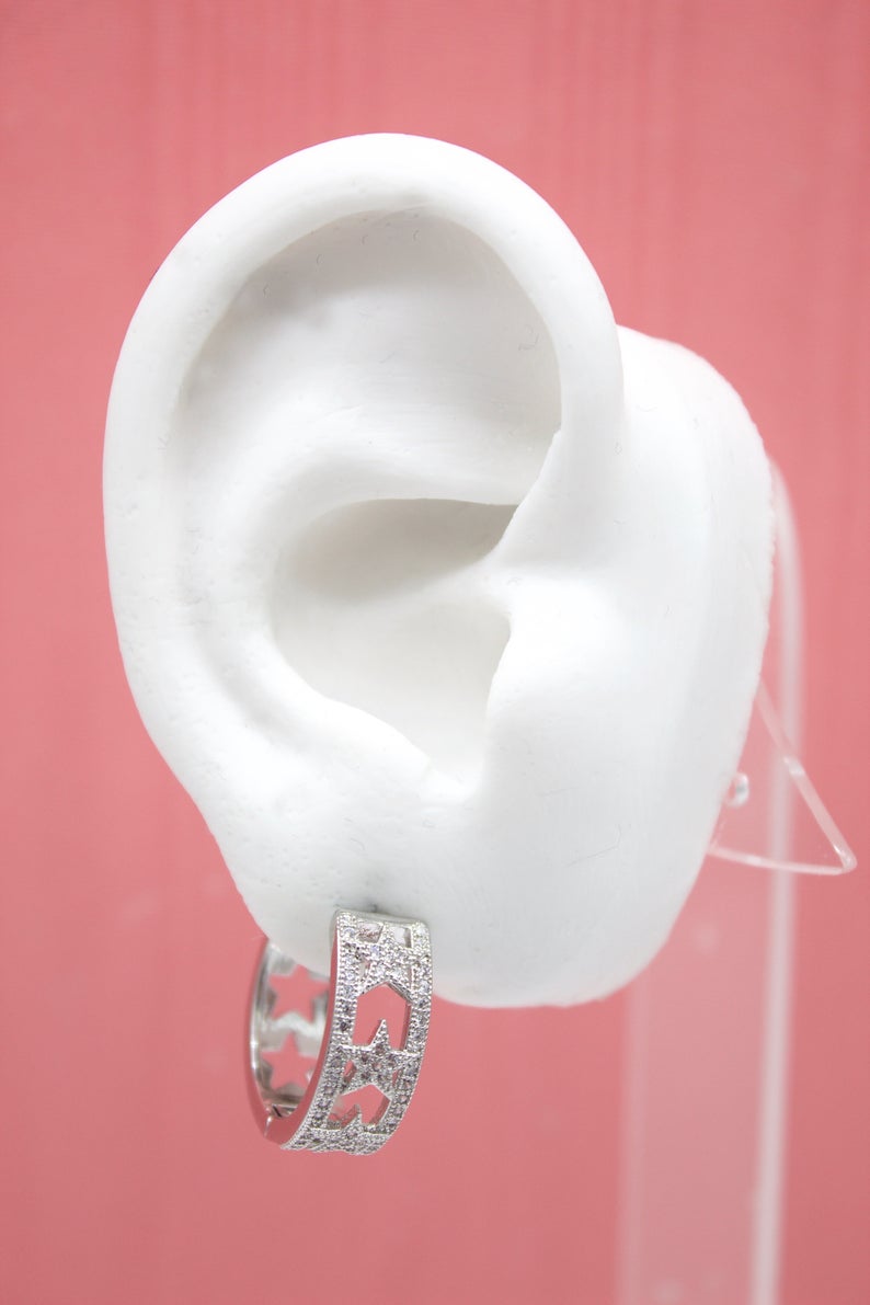 A white mannequin ear displays a sparkling silver huggie hoop earring adorned with cut-out star designs and pave stones against a pink background.