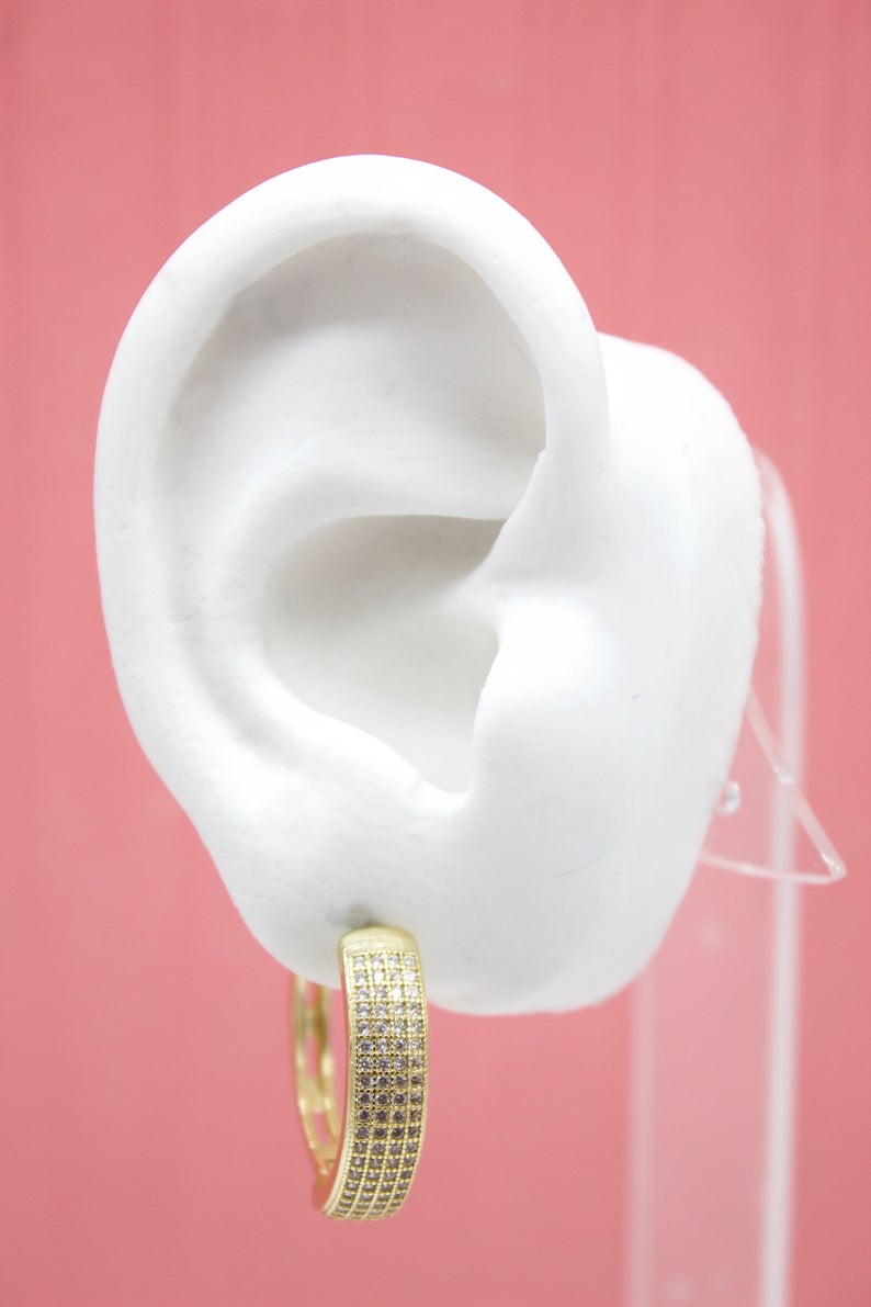 A white mannequin ear displays a paved gold hoop earring against a pink background.
