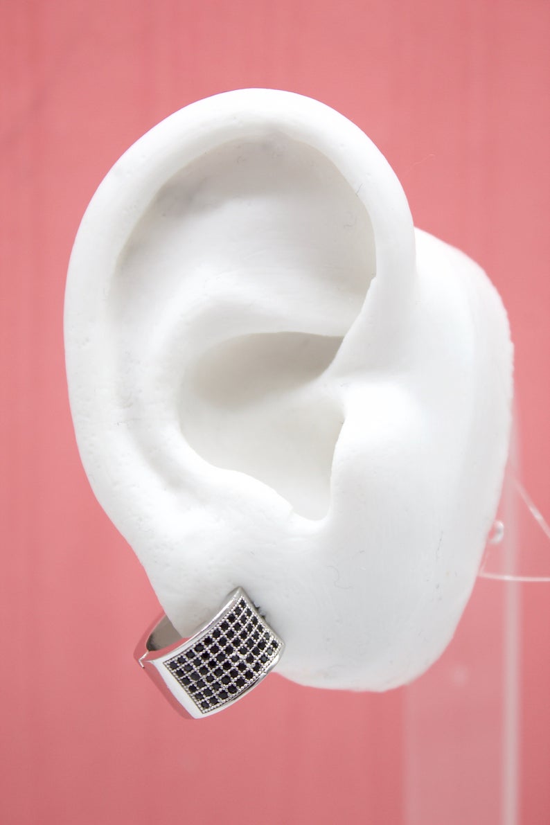 A white ear model displays a chunky silver hoop earring paved with tiny black cubic zirconia stones against a pink background.