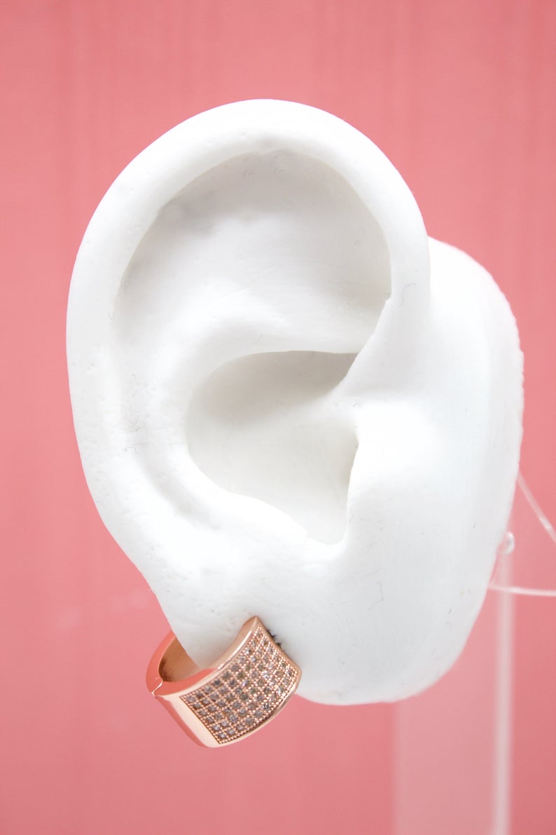 A white model ear displays a sparkling rose gold pave huggie hoop earring against a soft pink backdrop.