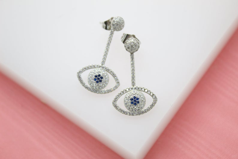 A pair of dazzling pave-set diamond evil eye drop earrings featuring sapphire blue centers rest against a white surface.