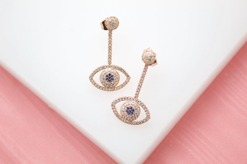 A pair of rose gold evil eye drop earrings embellished with sparkling white diamonds and deep blue sapphire centers rests on a white surface.