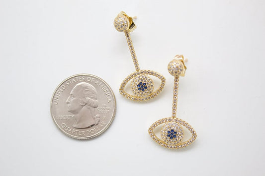 A pair of gold evil eye stud earrings adorned with clear and blue cubic zirconia rests beside a US quarter for scale.
