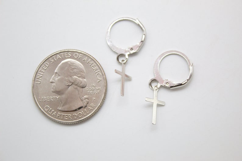 A pair of silver cross charm hoop earrings rests next to a US quarter coin for size comparison on a white surface.