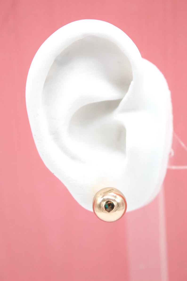 A glossy gold ball stud earring sparkles on a white mannequin ear against a solid pink background.