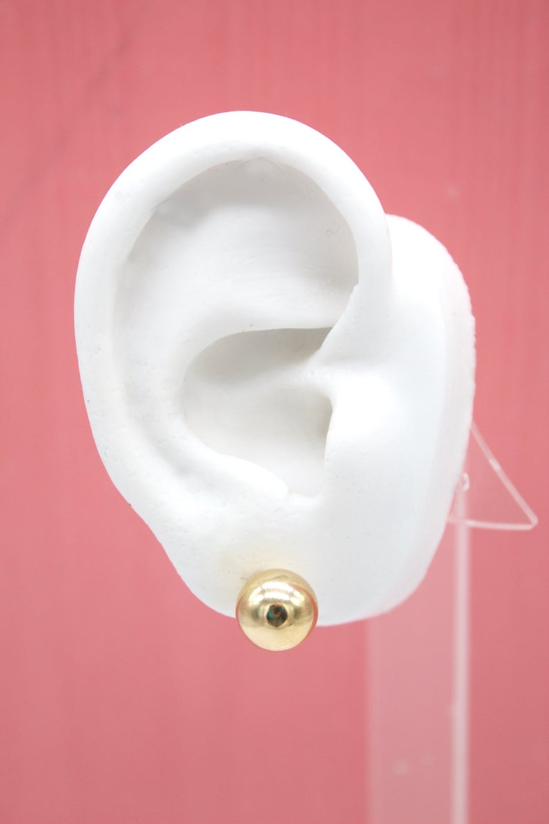 A white mannequin ear displays a polished gold ball stud earring against a pink background.