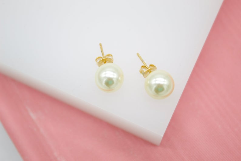 Two elegant gold faux pearl stud earrings rest on a white display block against a soft pink background.
