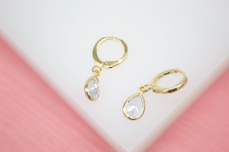 A pair of delicate gold hoop earrings with dangling teardrop-shaped crystal pendants rests on a white display surface.