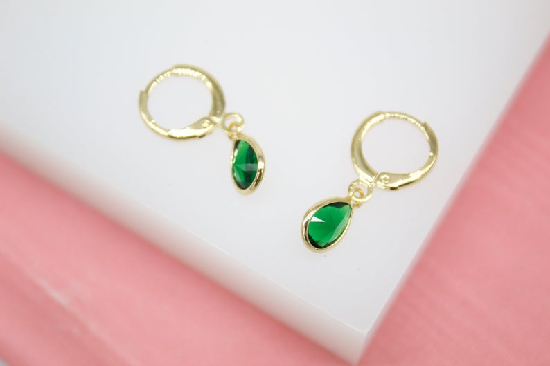 A pair of gold huggie hoop earrings featuring dangling teardrop green emerald crystals rests on a white display surface.