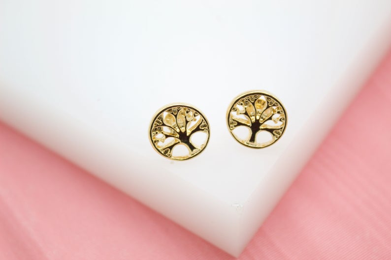 Two gold tree of life stud earrings rest on a white display block against a soft pink background.