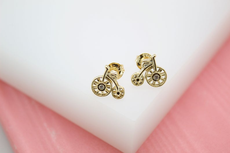 A pair of gold bicycle-shaped stud earrings featuring sparkling cubic zirconia stones rests on a white display block.