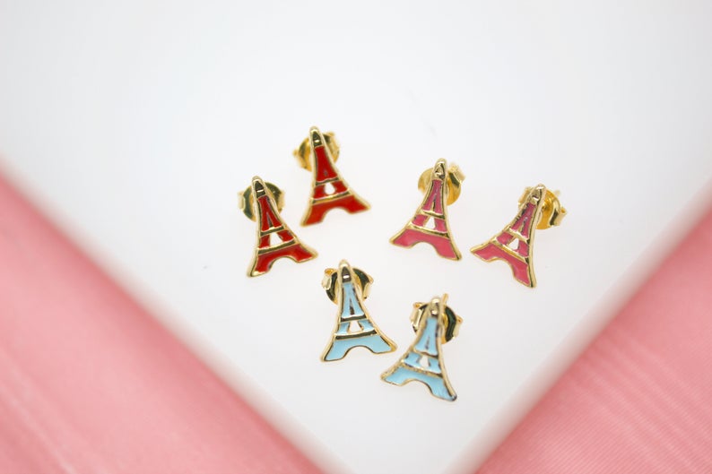 Six gold Eiffel Tower stud earrings in red, pink, and light blue enamel rest on a white surface.