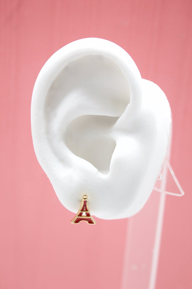 A white mannequin ear displays a gold and red Eiffel Tower stud earring against a pink background.