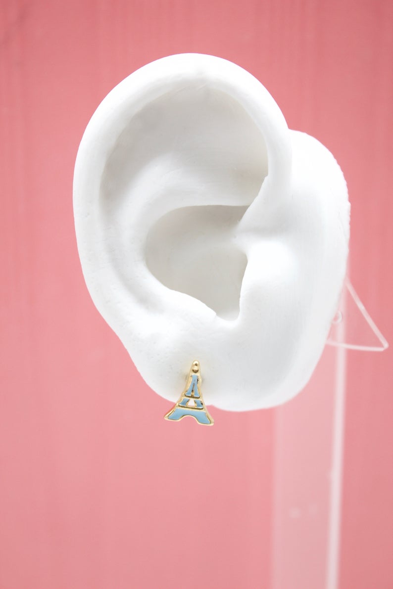 A white mannequin ear displays a gold and light blue enamel Eiffel Tower stud earring against a pink background.