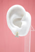 A white mannequin ear displays a gold and light blue enamel Eiffel Tower stud earring against a pink background.