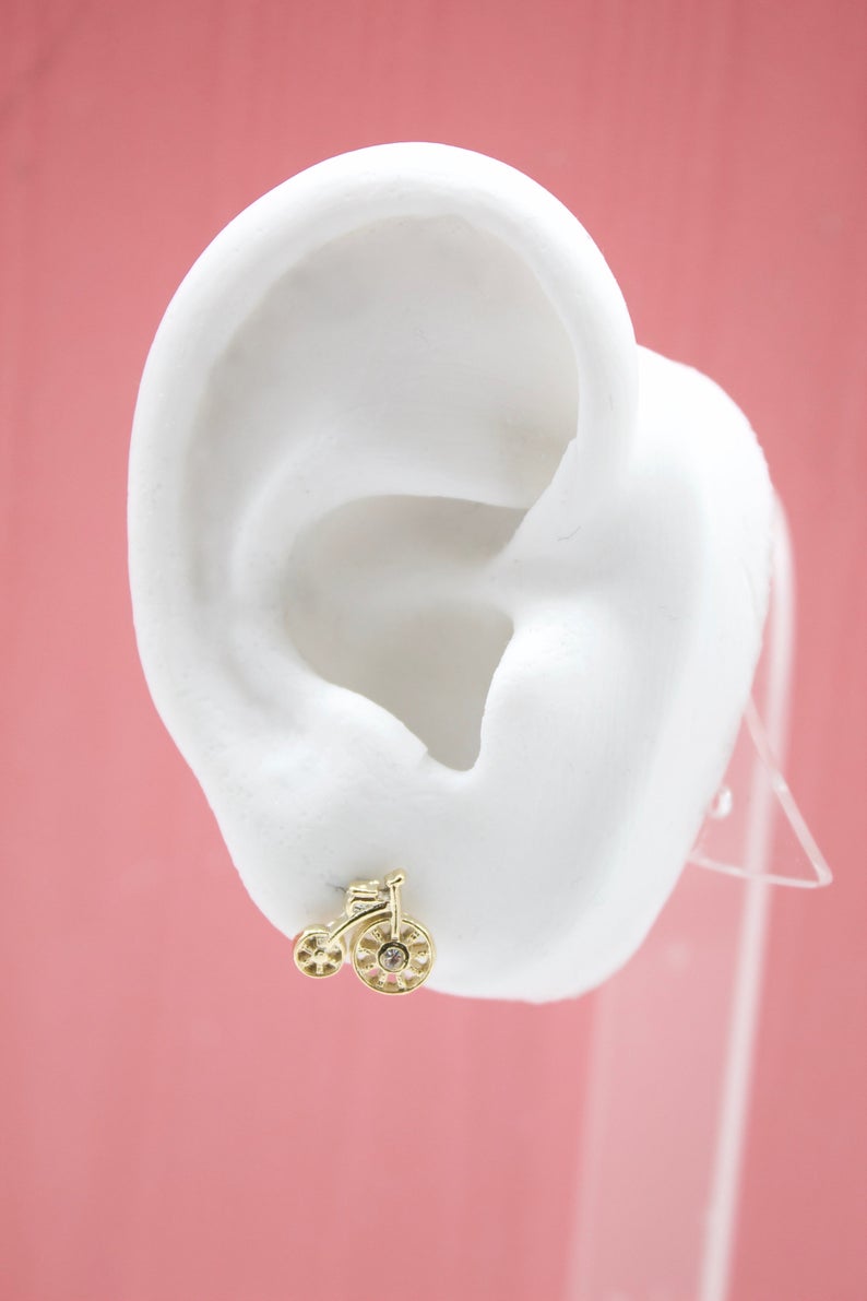 A cute gold bicycle stud earring sparkles on a white display ear against a pink background.