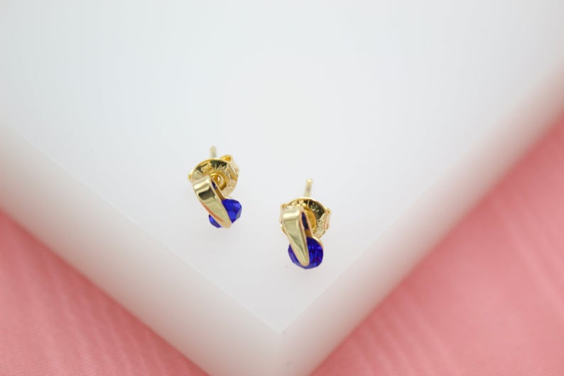 A pair of delicate gold huggie earrings featuring vibrant blue crystal stones rests on a white display surface.