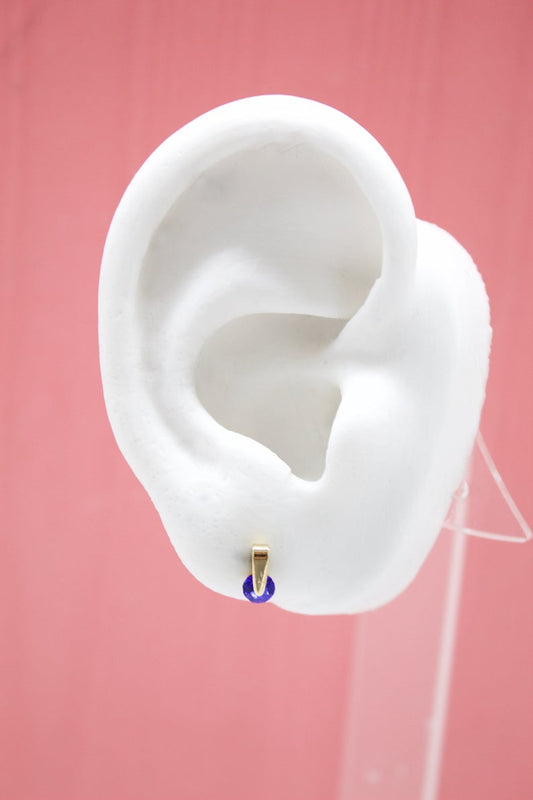 A white model ear displays a gold huggie hoop earring featuring a vibrant blue gemstone against a soft pink background.