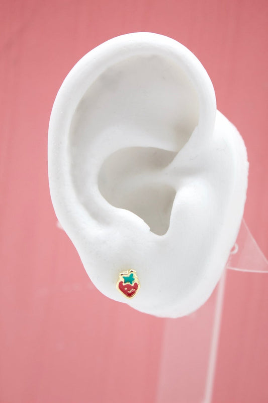 A white mannequin ear displays a tiny gold strawberry stud earring with red enamel and green leaves against a pink background.
