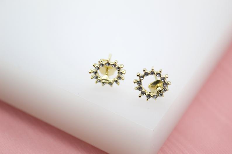 A pair of gold sunburst stud earrings encrusted with tiny cubic zirconia stones sits on a clean white display surface.