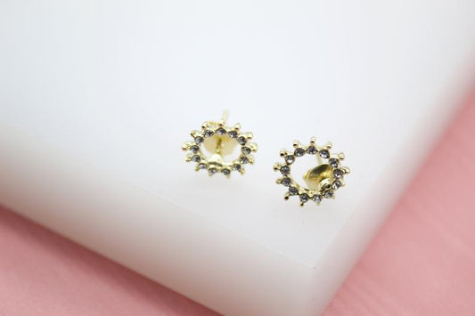 A pair of gold sunburst stud earrings encrusted with tiny cubic zirconia stones sits on a clean white display surface.