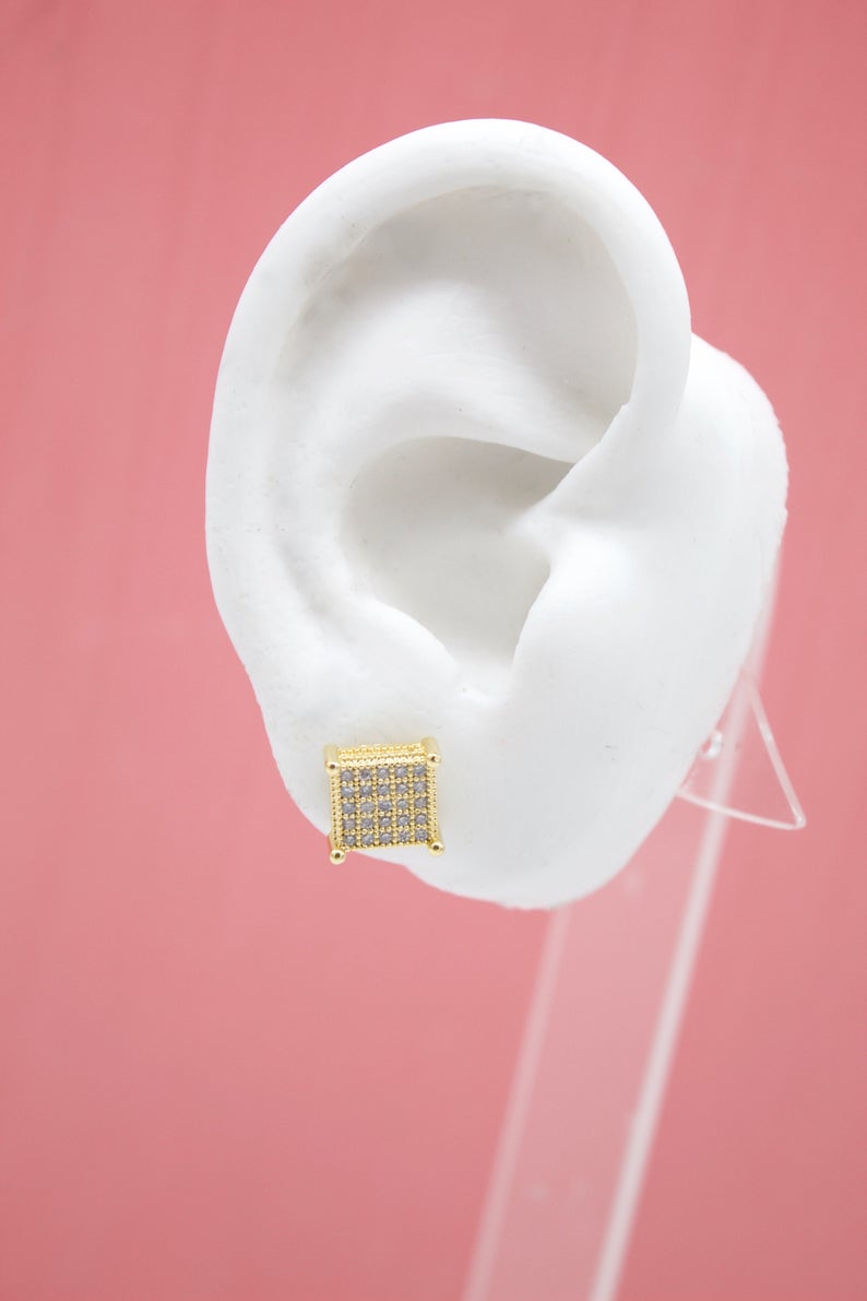 A white model ear displays a dazzling square gold stud earring encrusted with sparkling pave diamonds against a solid pink backdrop.