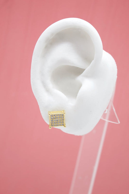 A white model ear displays a dazzling square gold stud earring encrusted with sparkling pave diamonds against a solid pink backdrop.