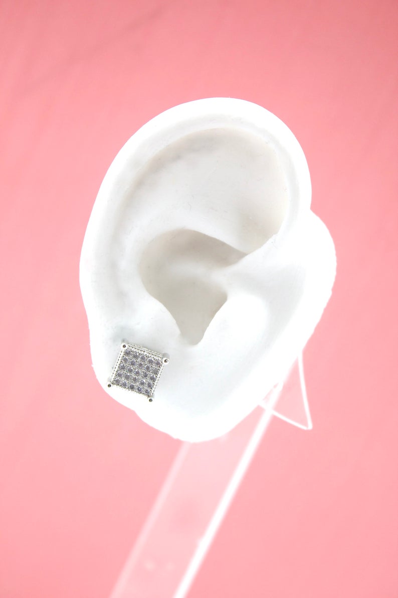 A white mannequin ear on a pink background displays a sparkling square stud earring.