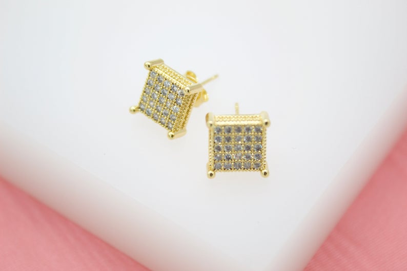 A pair of gold square stud earrings encrusted with sparkling micro pavé stones rests on a white display surface.