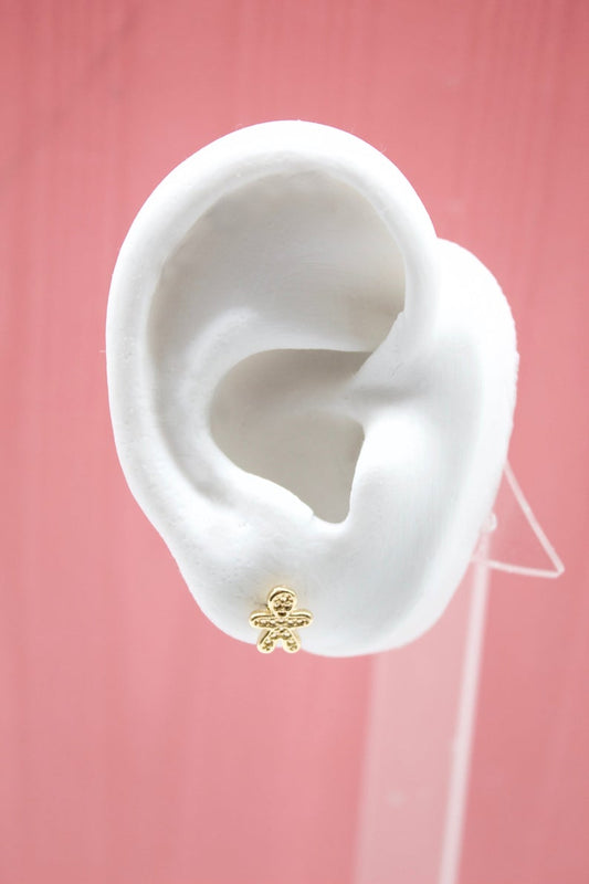 A white mannequin ear displays a cute gold gingerbread man stud earring against a pink background.