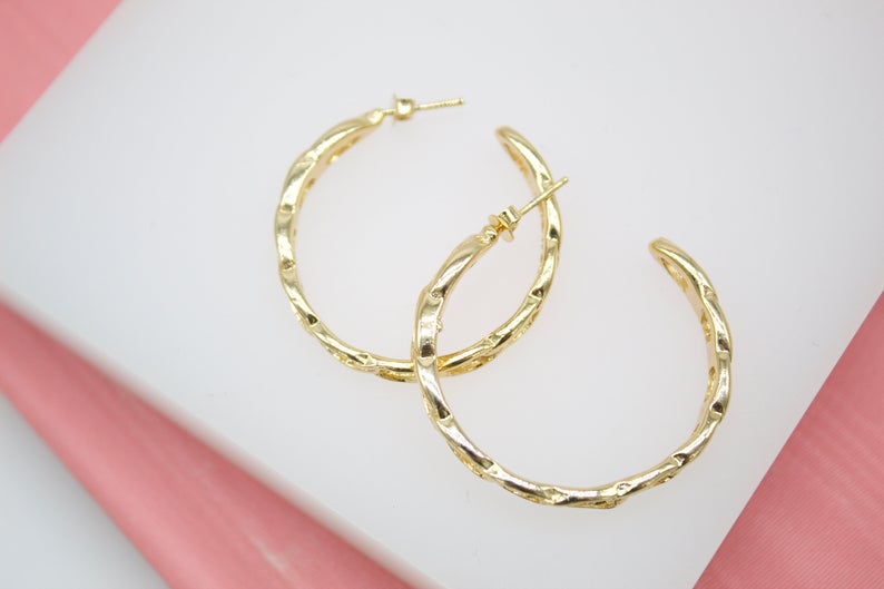 Two textured gold hoop earrings rest on a white display surface against a pink background.