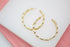 Two textured gold hoop earrings rest on a white display surface against a pink background.