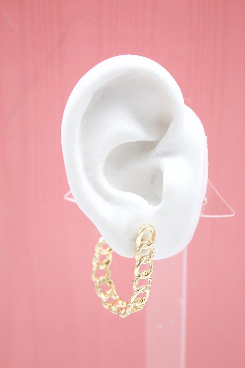 A white mannequin ear displays a textured gold chain-link hoop earring against a pink background.
