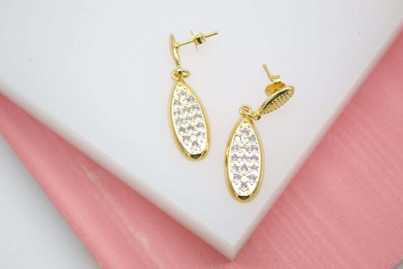 A pair of gold drop earrings encrusted with sparkling cubic zirconia stones rests on a white display surface.