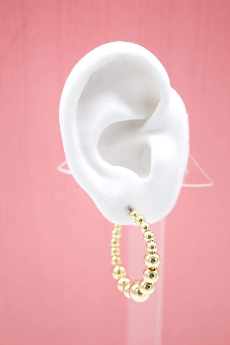 A white mannequin ear displays a graduated gold bead hoop earring against a pink background.