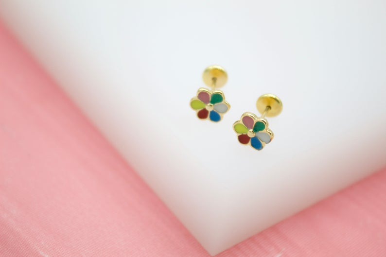 A pair of gold flower stud earrings featuring colorful enamel petals rests on a white display surface against a pink background.