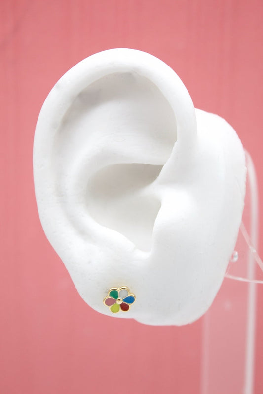 A white model ear displays a gold flower stud earring featuring multicolored enamel petals against a solid pink background.