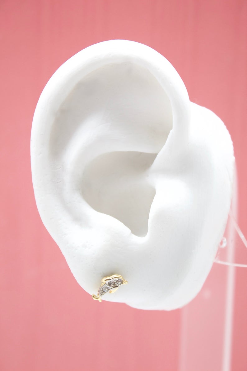 A white mannequin ear displays a sparkling gold dolphin stud earring against a soft pink background.