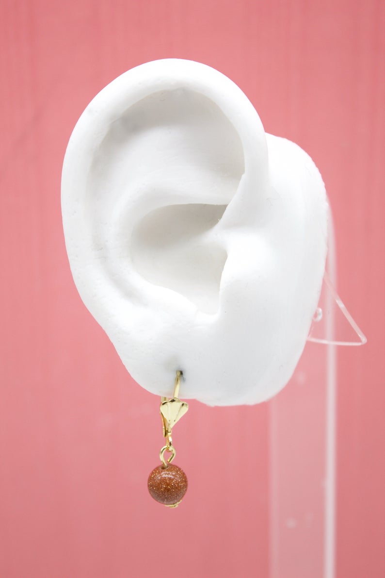 A white mannequin ear displays a gold-plated leverback earring featuring a sparkling brown goldstone gemstone bead.