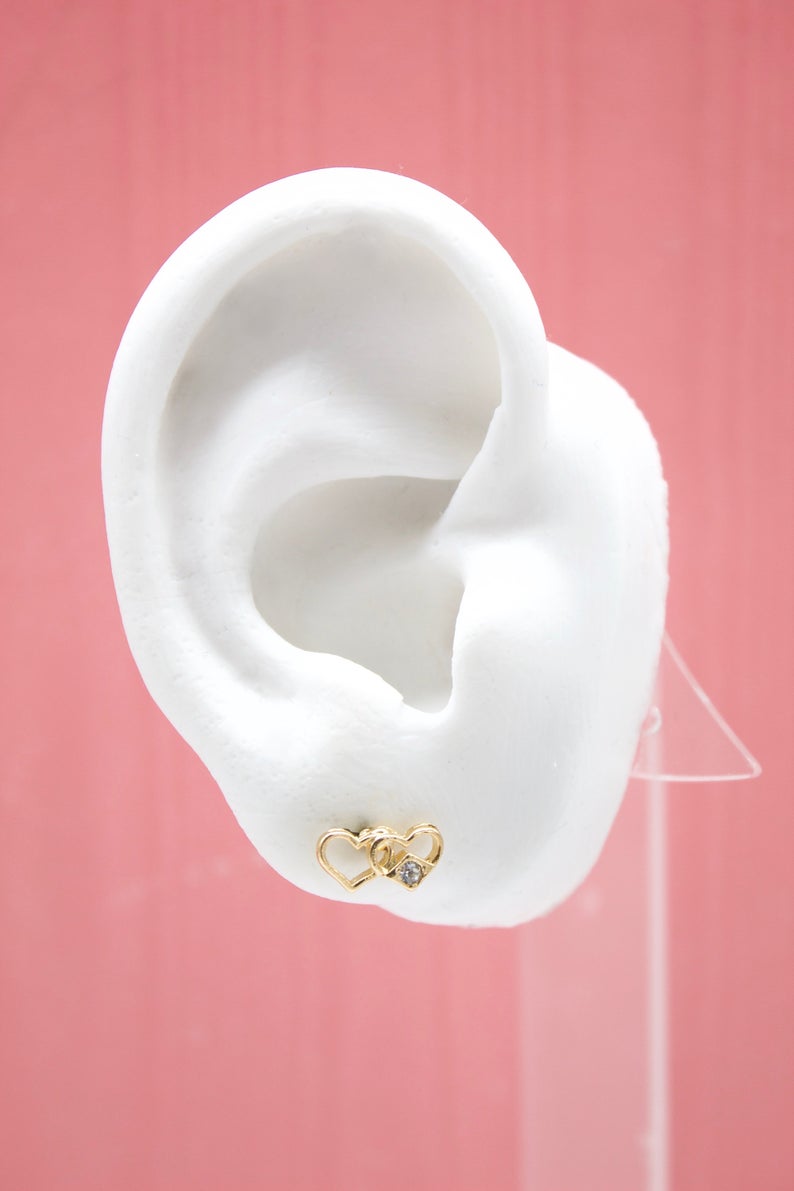 A white mannequin ear displays a gold double heart stud earring with a tiny sparkling diamond.