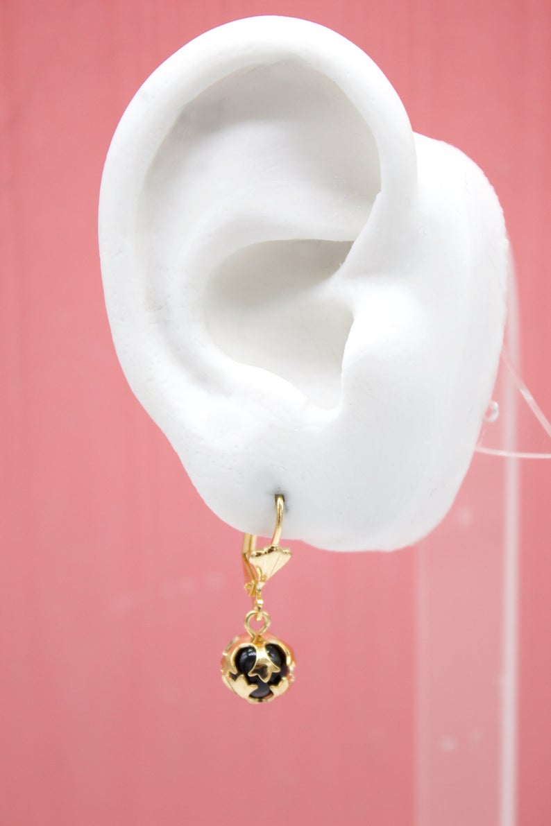 A white mannequin ear displays a gold leverback earring featuring a black bead encased in a gold filigree cage.