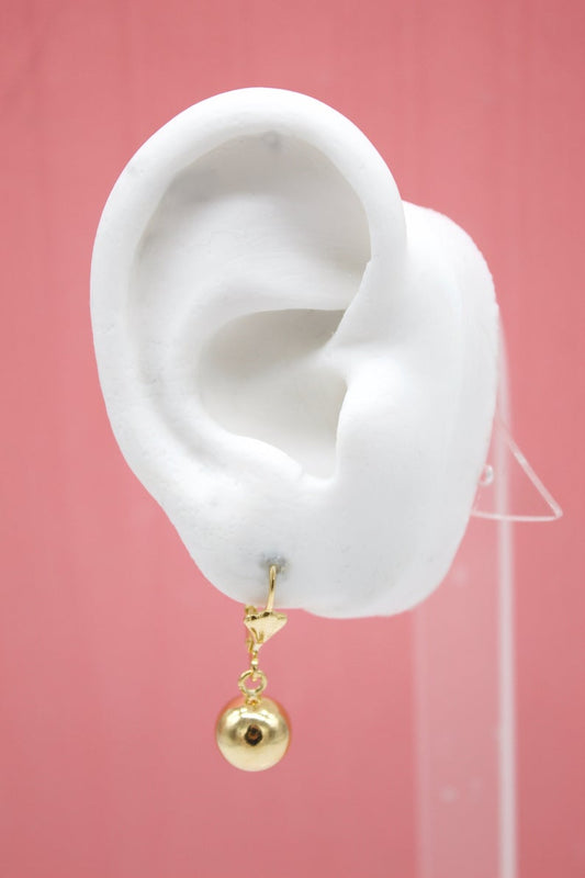 A white mannequin ear displays a dangling gold ball charm earring against a pink background.
