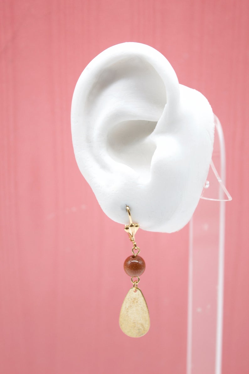 A white mannequin ear displays a dangling gold earring featuring a sparkling brown gemstone and a teardrop-shaped metal charm.