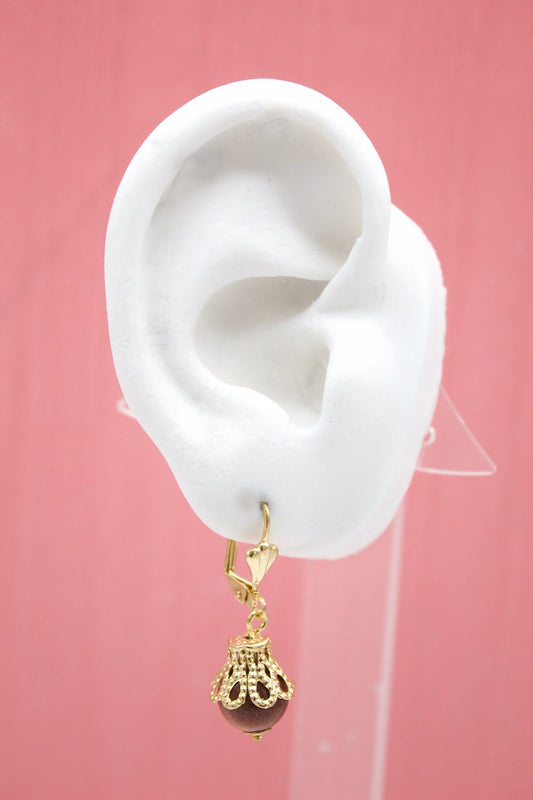 A white mannequin ear displays a dangling gold filigree leverback earring featuring a round brown gemstone against a pink background.