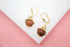 A pair of gold leverback earrings featuring dangling spherical brown gemstone beads rests on a white display surface.