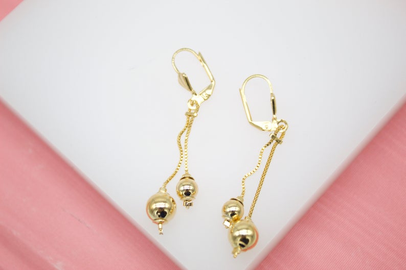 A pair of gold dangle earrings featuring delicate chains and polished sphere beads rests on a white display surface.